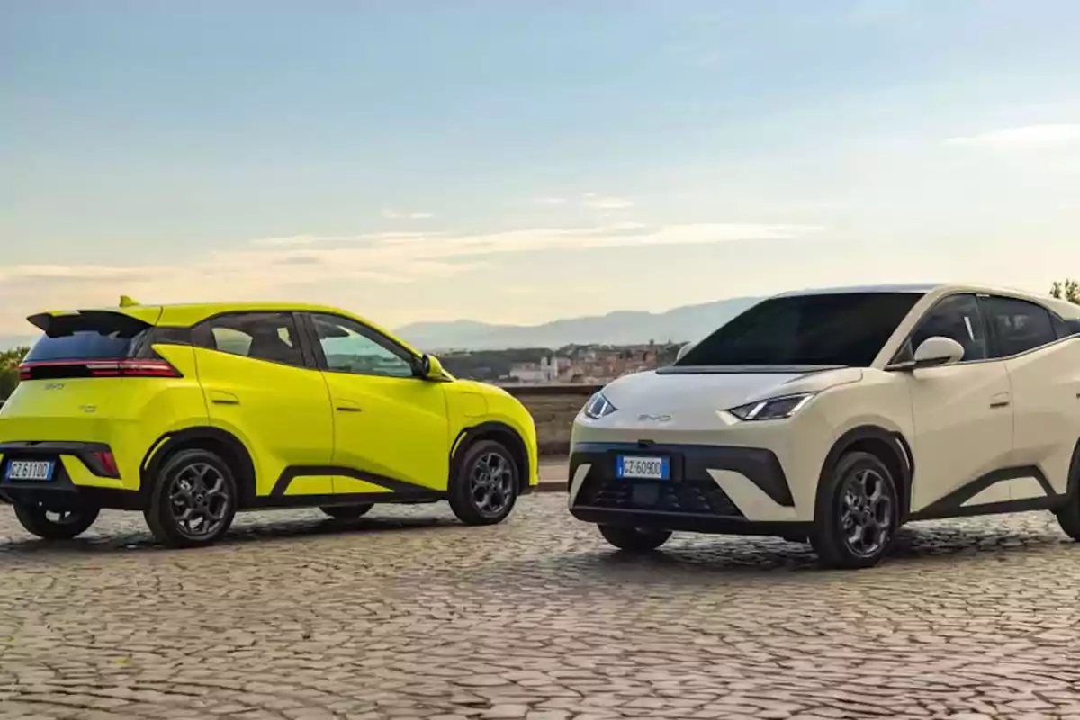 Dos autos eléctricos de colores amarillo y blanco estacionados sobre un camino de adoquines con un paisaje urbano y montañas al fondo. Dos autos eléctricos de colores amarillo y blanco estacionados sobre un camino de adoquines con un paisaje urbano y montañas al fondo.