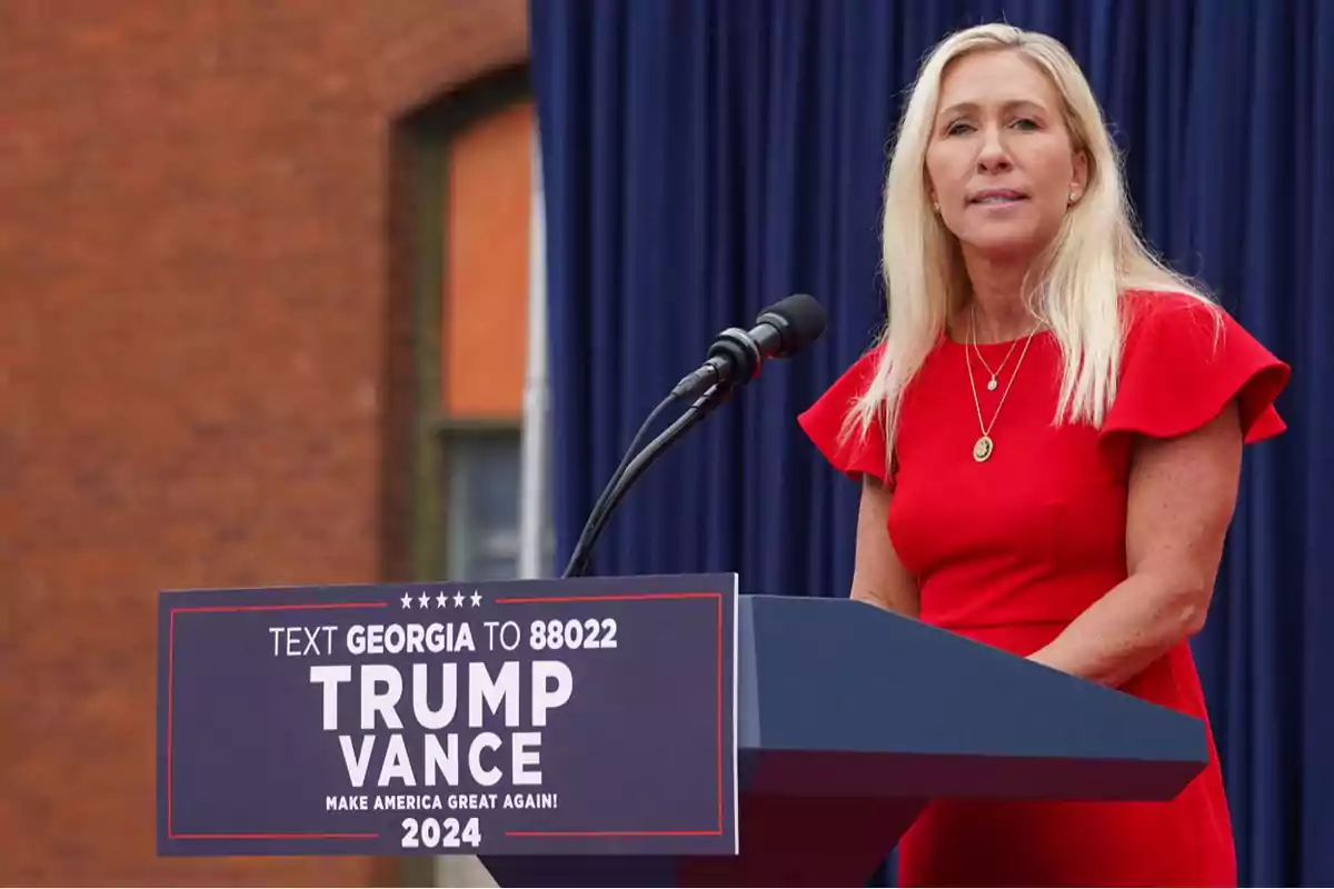 A blonde-haired woman in a red dress speaks at a podium with a sign that says A blonde-haired woman in a red dress speaks at a podium with a sign that says