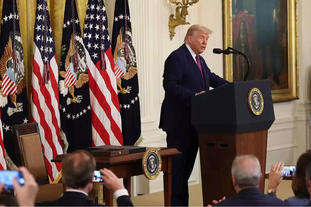 A man in a dark suit speaks at a presidential podium in a room decorated with American flags while several people take his picture.