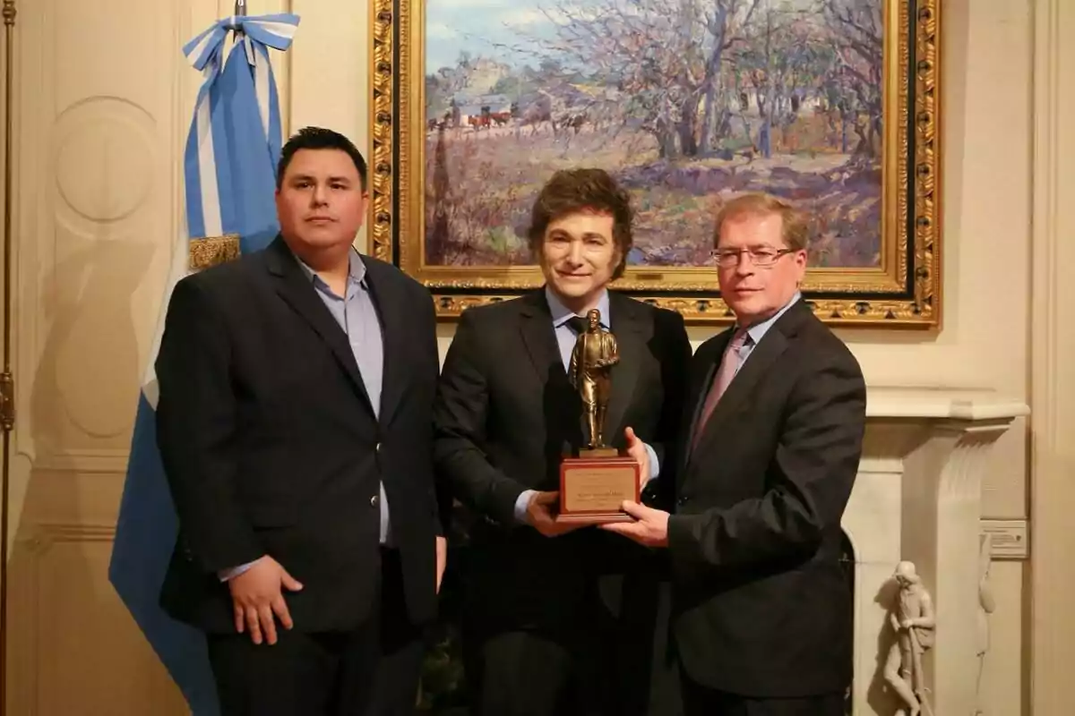 Tres hombres de traje posan con una estatua en un interior elegante con una bandera argentina y un cuadro de paisaje al fondo.