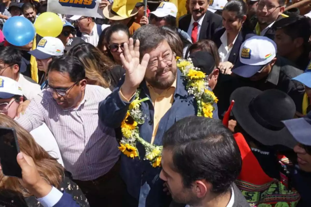 Un hombre con gafas y una guirnalda de flores amarillas saluda mientras está rodeado de una multitud de personas, algunas con gorras y globos. Un hombre con gafas y una guirnalda de flores amarillas saluda mientras está rodeado de una multitud de personas, algunas con gorras y globos.