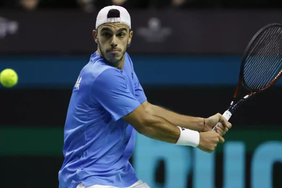 Un tenista con camiseta azul y gorra blanca se prepara para golpear la pelota con su raqueta.