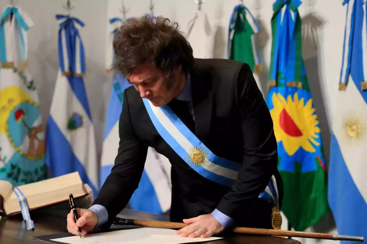 Un hombre con una banda presidencial firma un documento en una mesa, rodeado de banderas argentinas.