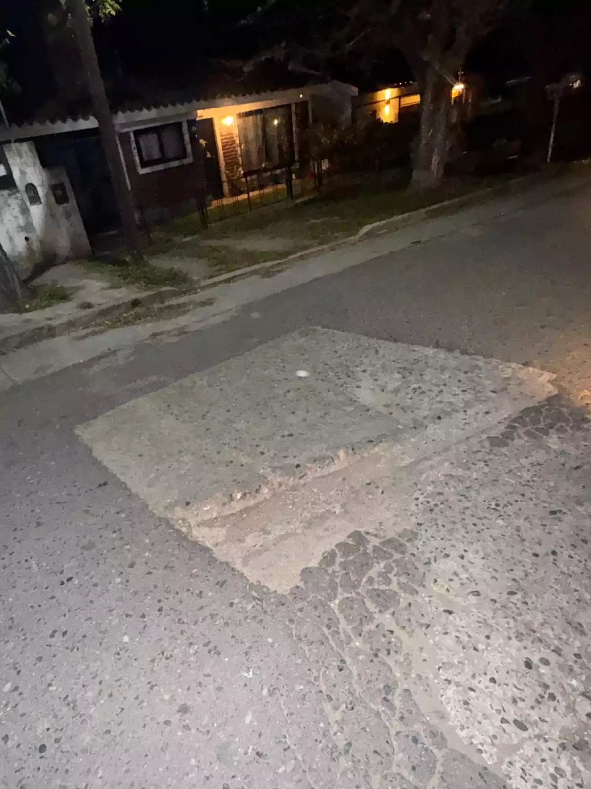 A street with a large, deep pothole in the pavement, illuminated by the lights of nearby houses at night. A street with a large, deep pothole in the pavement, illuminated by the lights of nearby houses at night.