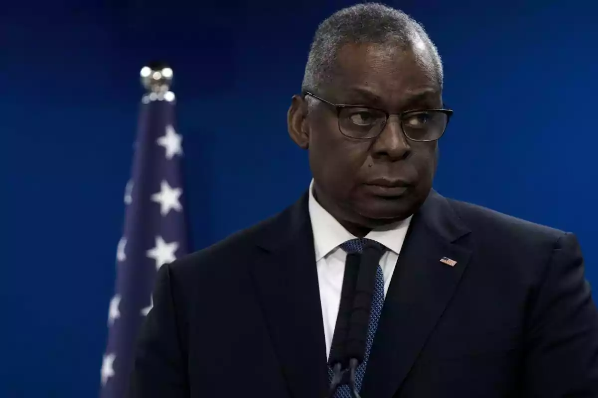 Hombre de traje oscuro y gafas frente a un micrófono con una bandera de fondo.