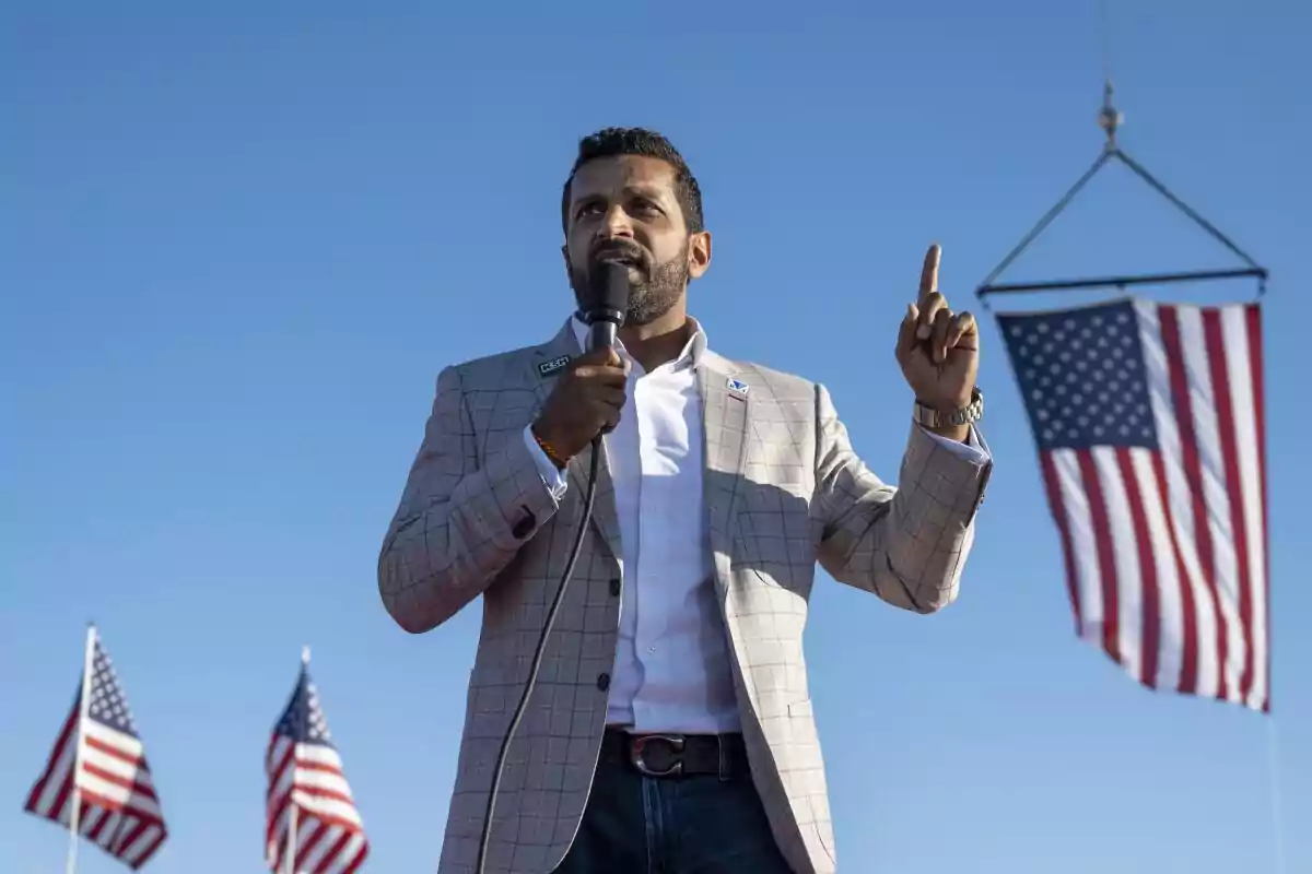 Man speaking at an outdoor rally with American flags in the background.