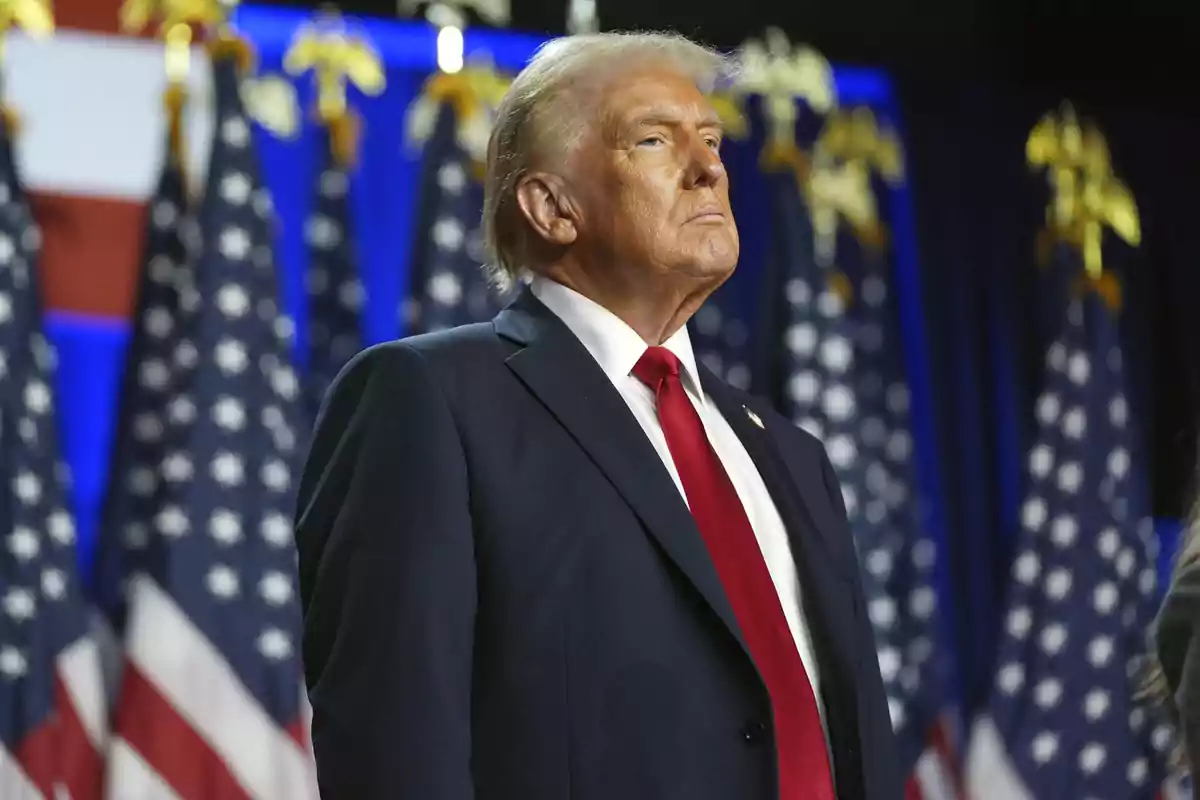 Hombre de cabello rubio con traje oscuro y corbata roja de pie frente a varias banderas de Estados Unidos Hombre de cabello rubio con traje oscuro y corbata roja de pie frente a varias banderas de Estados Unidos