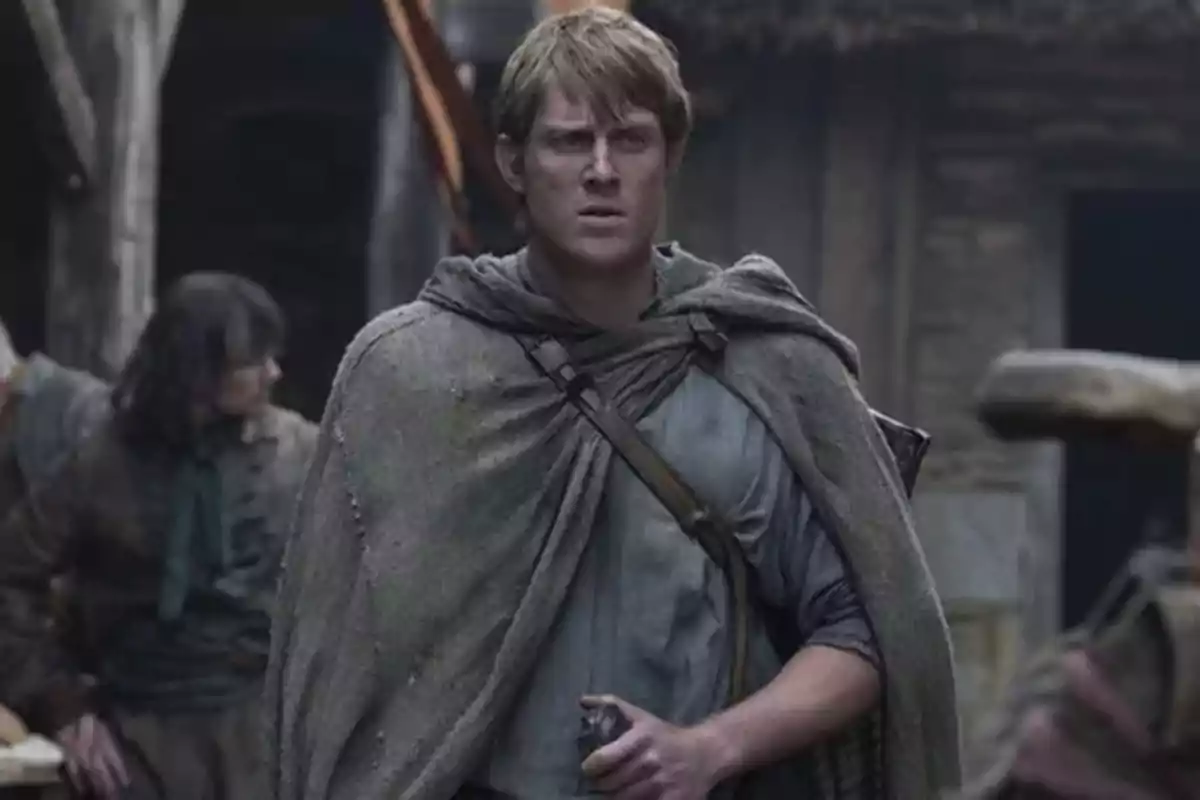 Un hombre con capa gris y expresión seria está de pie en un entorno medieval con edificios de madera y personas al fondo. Un hombre con capa gris y expresión seria está de pie en un entorno medieval con edificios de madera y personas al fondo.