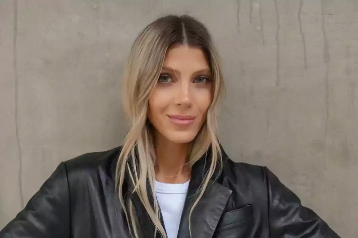 Mujer rubia con chaqueta de cuero negro y camiseta blanca posando frente a una pared de concreto