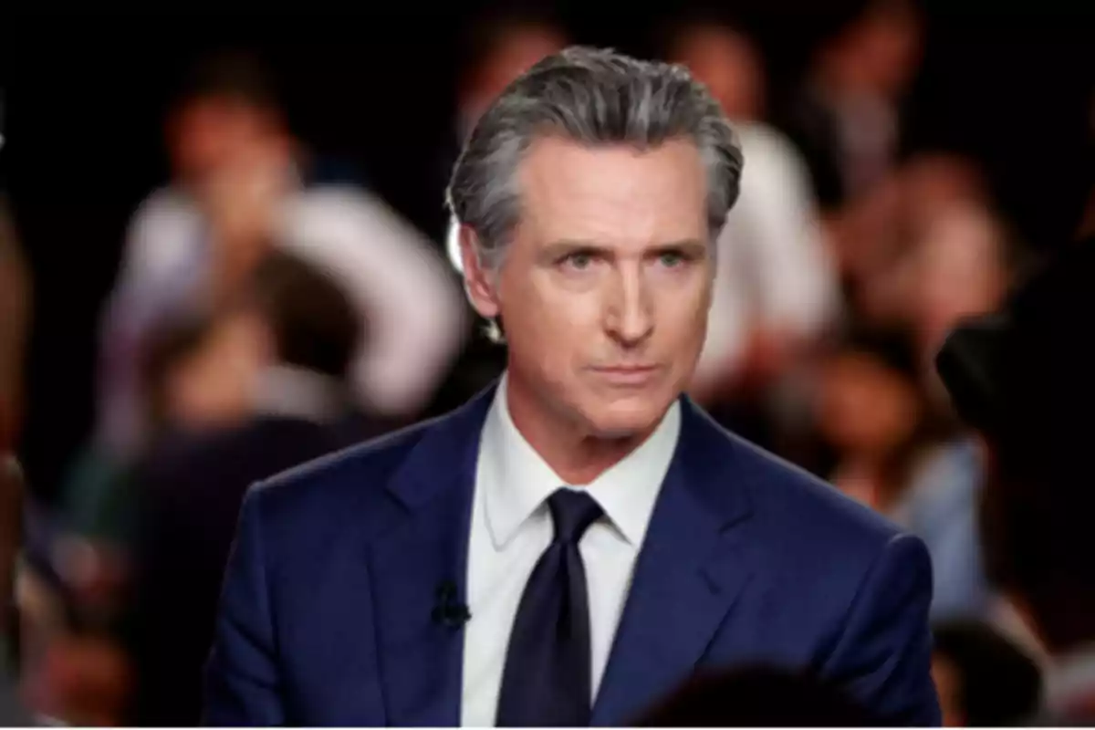 Hombre de cabello canoso con traje azul y corbata oscura en un entorno con personas desenfocadas al fondo Hombre de cabello canoso con traje azul y corbata oscura en un entorno con personas desenfocadas al fondo