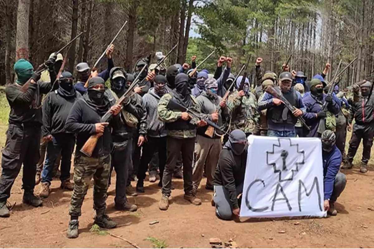 Un grupo de personas enmascaradas y armadas posan en un bosque sosteniendo un cartel que dice "CAM".