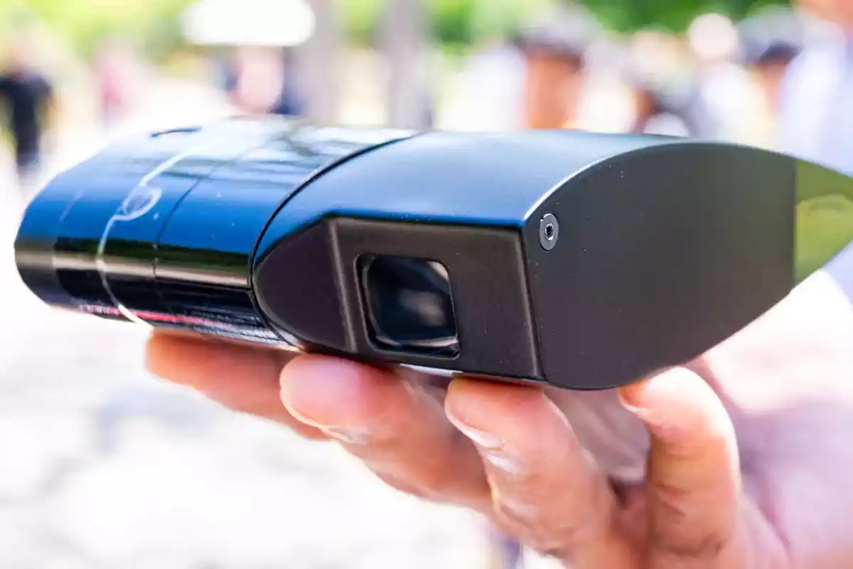 Persona sosteniendo un dispositivo electrónico negro en la mano con fondo desenfocado al aire libre Persona sosteniendo un dispositivo electrónico negro en la mano con fondo desenfocado al aire libre