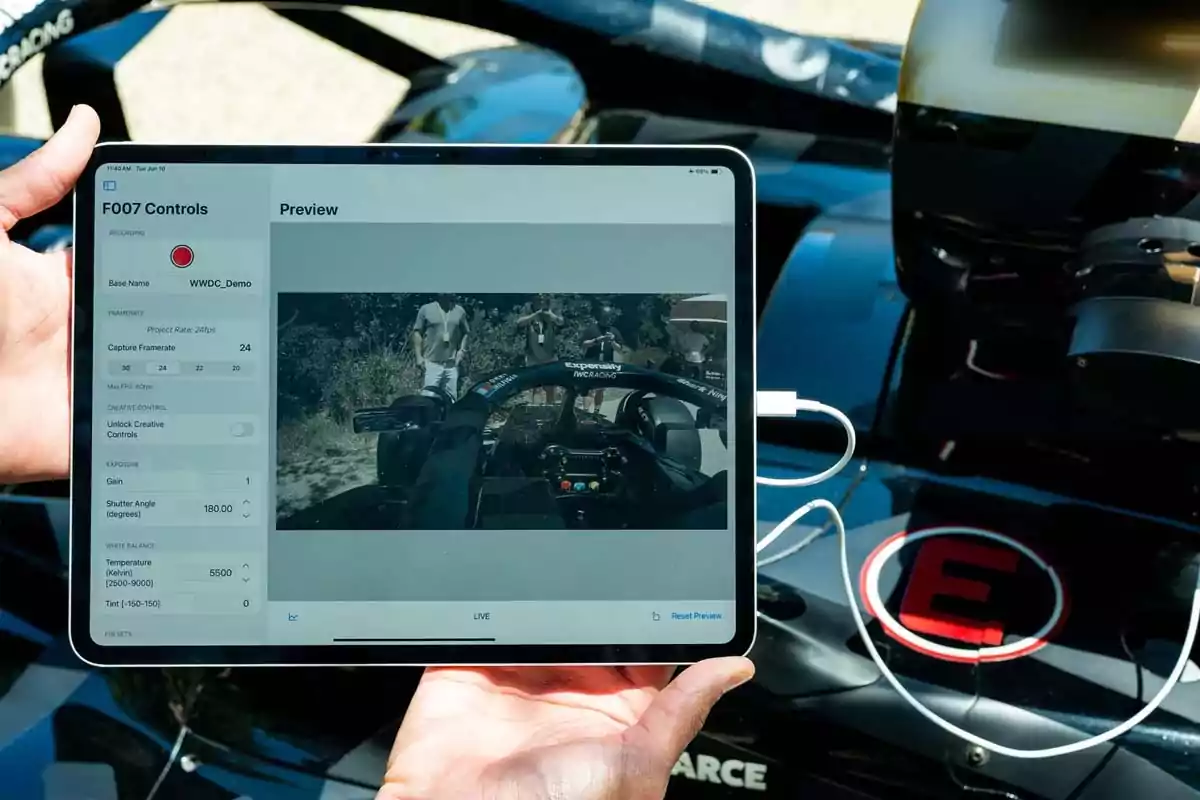 Una persona sostiene una tablet que muestra una transmisión en vivo desde el interior de un auto de carreras, con controles de grabación visibles en la pantalla y el vehículo de fondo. Una persona sostiene una tablet que muestra una transmisión en vivo desde el interior de un auto de carreras, con controles de grabación visibles en la pantalla y el vehículo de fondo.