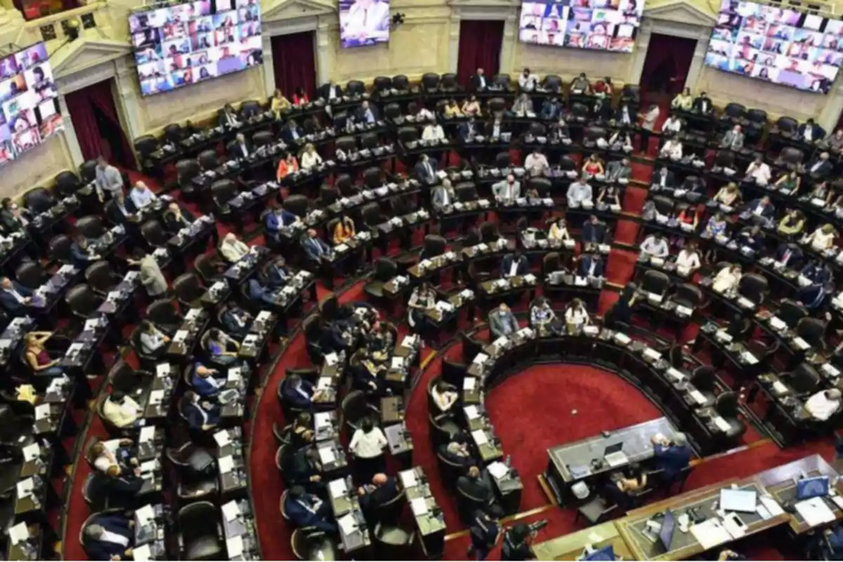 Una sala de asamblea con numerosos asientos ocupados y pantallas mostrando videollamadas.