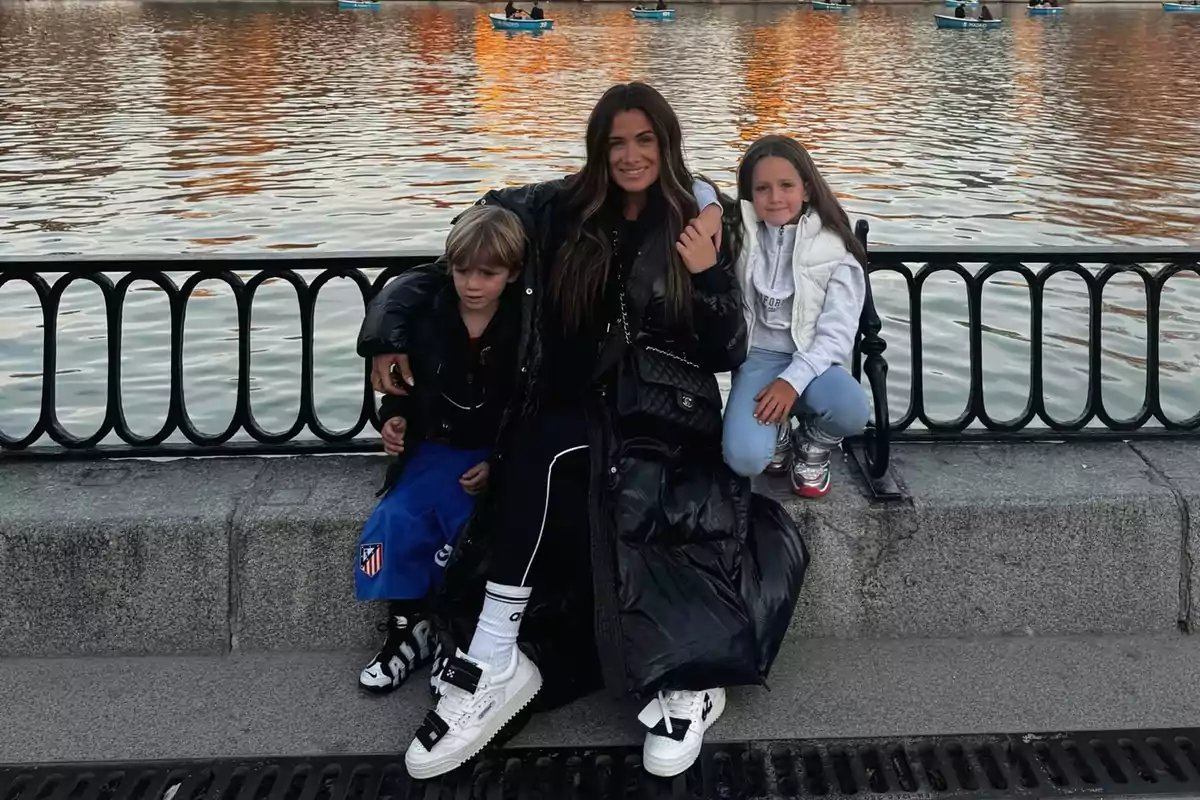 Una mujer y dos niños posan juntos sentados en un muro frente a un lago con botes al fondo. Una mujer y dos niños posan juntos sentados en un muro frente a un lago con botes al fondo.