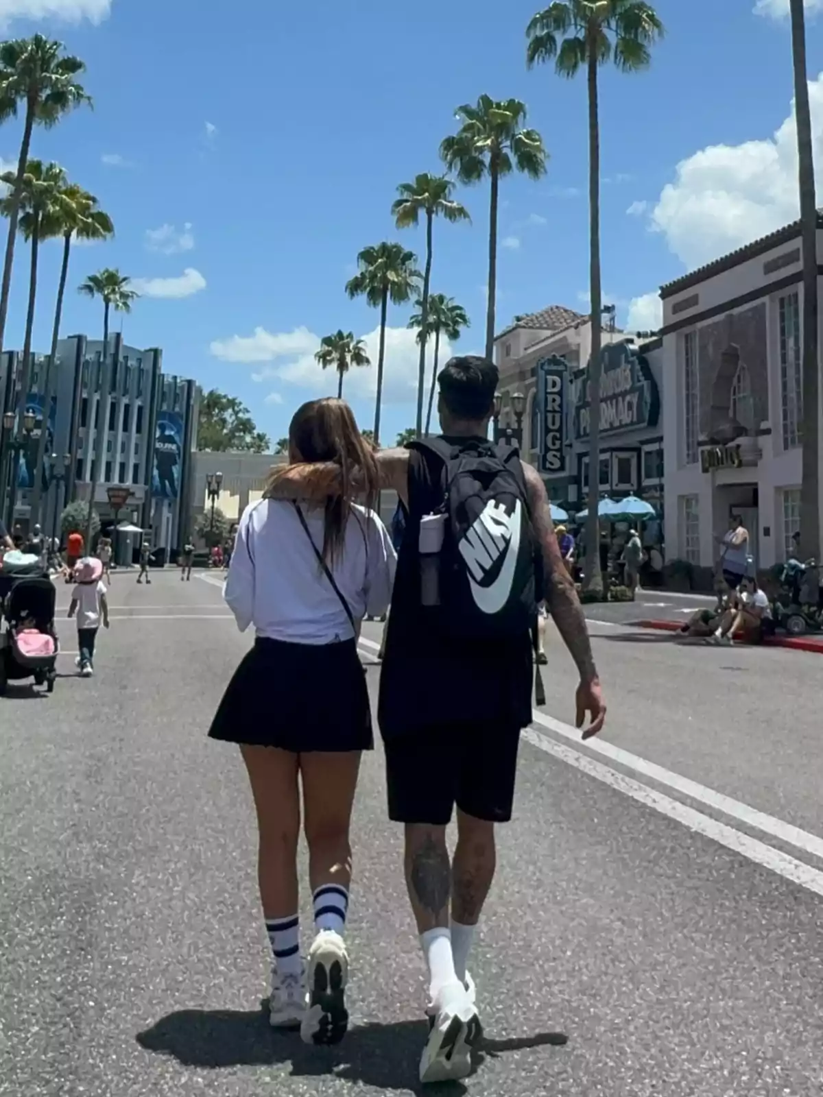 Pareja caminando abrazada por una avenida soleada con palmeras y edificios al fondo Pareja caminando abrazada por una avenida soleada con palmeras y edificios al fondo