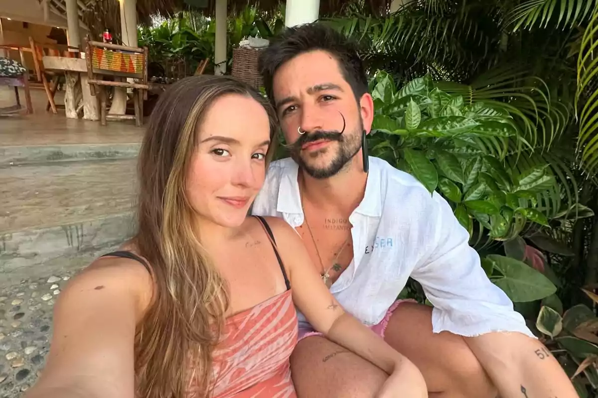 Una pareja sonríe mientras se toma una selfie en un entorno tropical con plantas y muebles de madera al fondo.