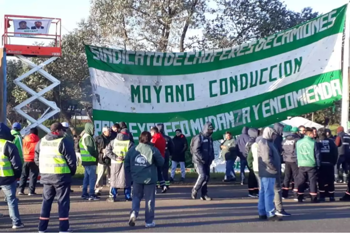 Un grupo de personas se encuentra reunido frente a una gran pancarta verde y blanca que dice 