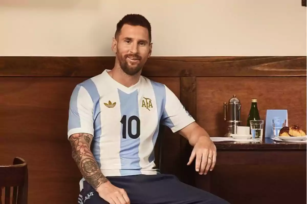 Un hombre con camiseta de fútbol celeste y blanca sentado en un café con una mesa que tiene una botella, un vaso de agua y un plato con medialunas.