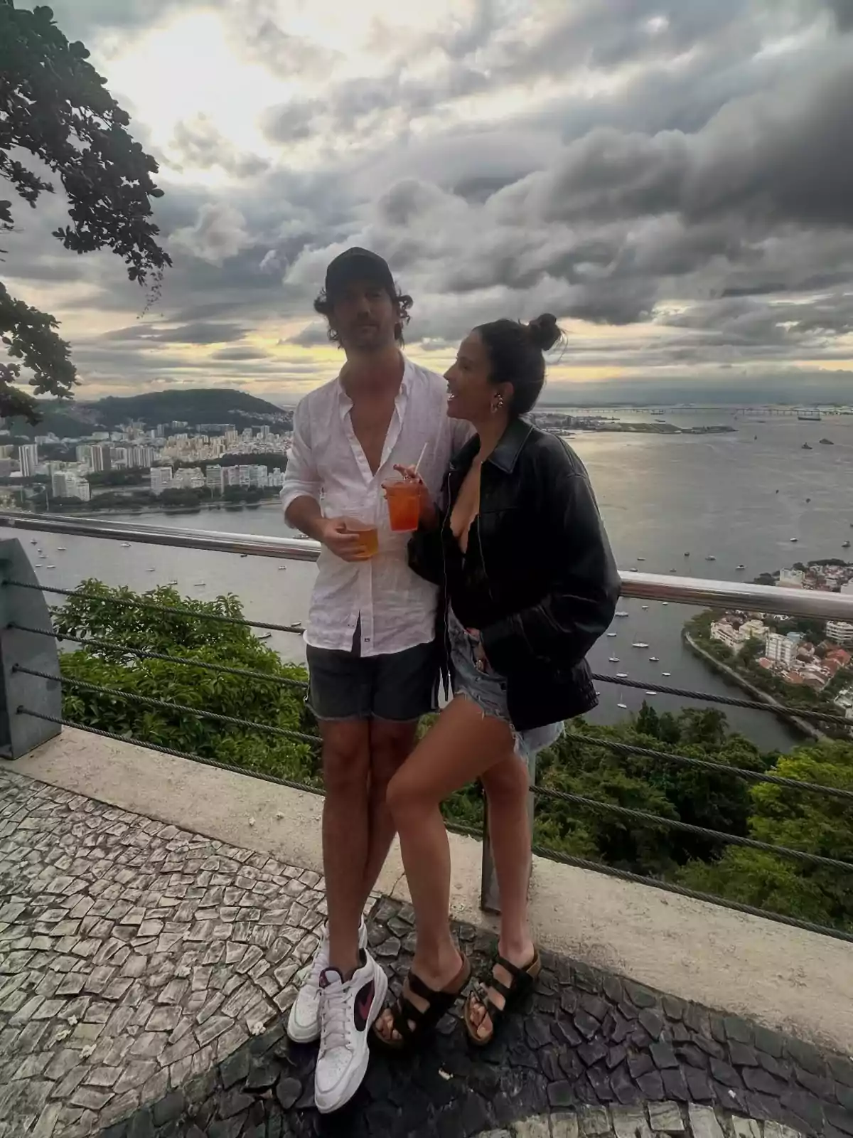 Pareja disfrutando de bebidas al aire libre con vista panorámica a una ciudad y un río bajo un cielo nublado Pareja disfrutando de bebidas al aire libre con vista panorámica a una ciudad y un río bajo un cielo nublado