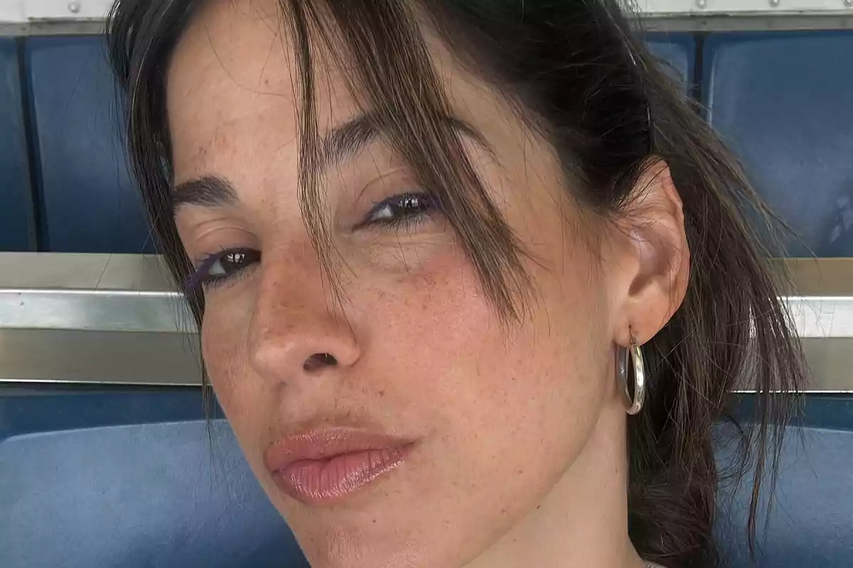 Mujer de cabello castaño con flequillo y arete de aro plateado mirando a la cámara en un lugar con asientos azules
