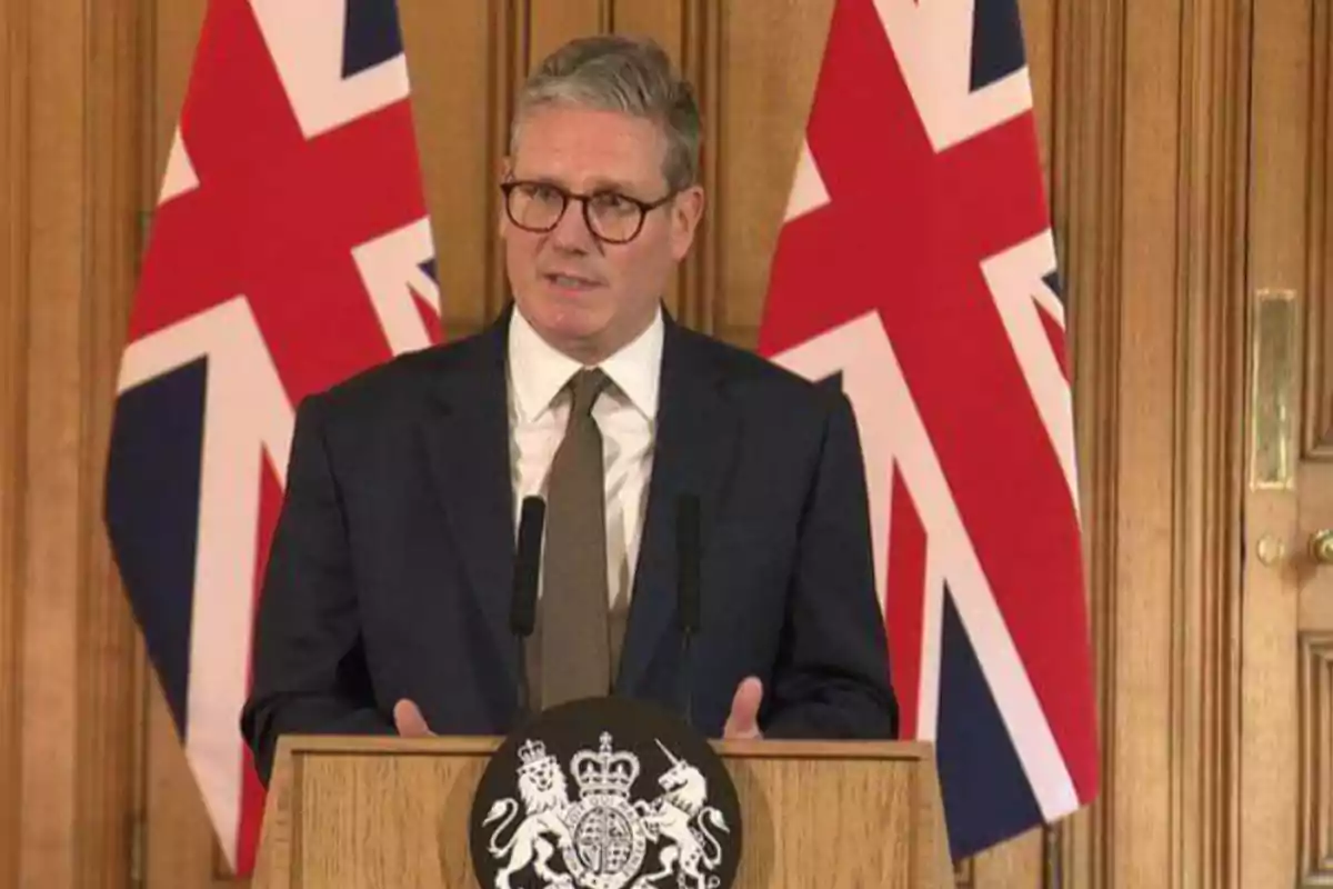 Un hombre con gafas y traje oscuro hablando en un podio con el escudo del Reino Unido, con dos banderas británicas de fondo.