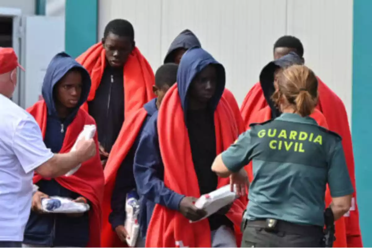 Un grupo de personas con mantas rojas y capuchas recibe ayuda de un miembro de la Guardia Civil y otro individuo.