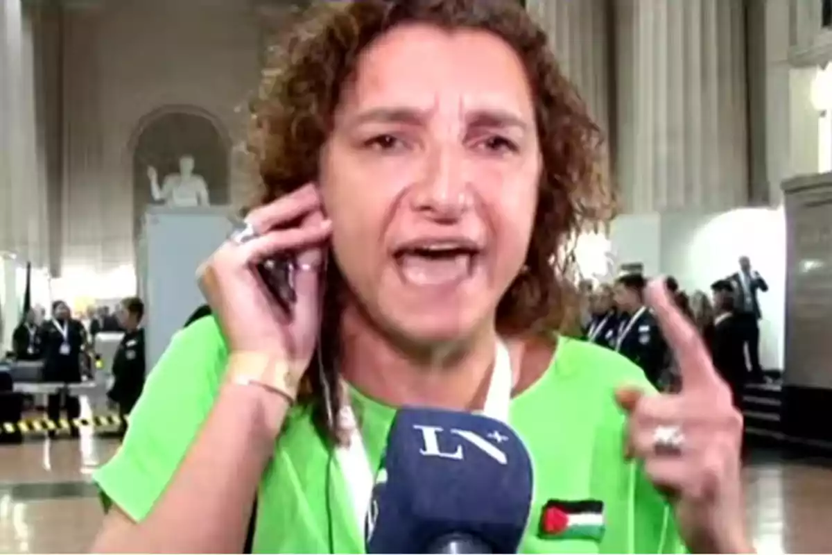 Una mujer con una camiseta verde habla en un micrófono mientras sostiene un teléfono móvil en una mano y gesticula con la otra, en un entorno interior con varias personas al fondo.