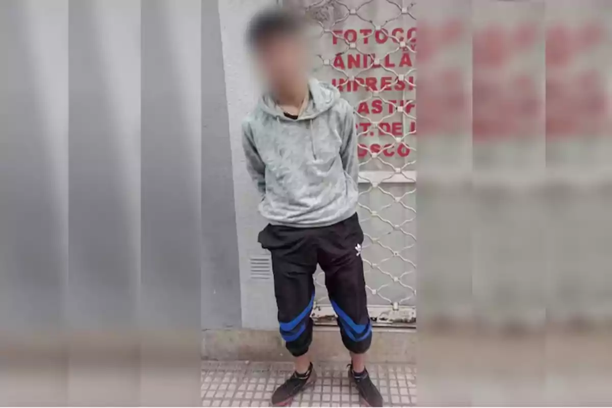 Una persona con el rostro difuminado está de pie frente a una reja metálica, vistiendo una sudadera gris y pantalones deportivos negros con detalles azules.