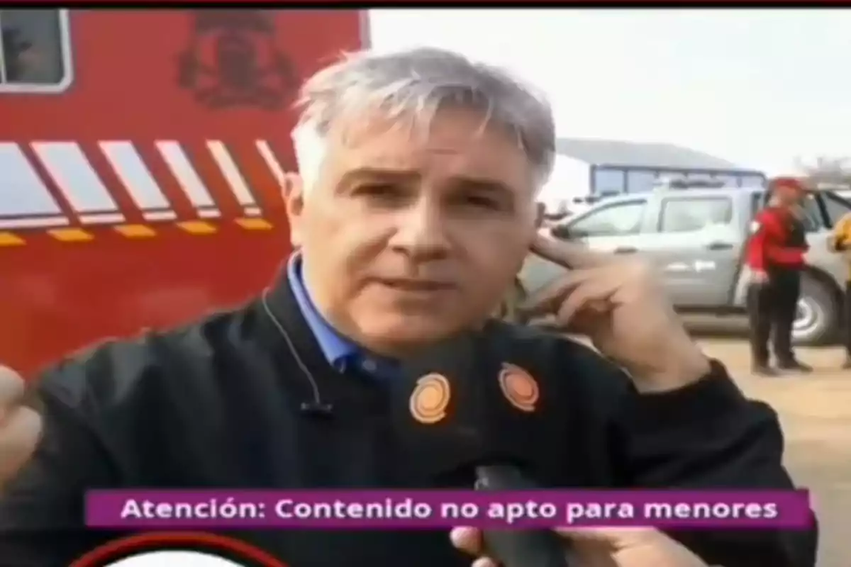 Un hombre con cabello canoso está siendo entrevistado frente a un vehículo de emergencia, mientras en la parte inferior de la imagen se muestra un aviso que dice "Atención: Contenido no apto para menores".