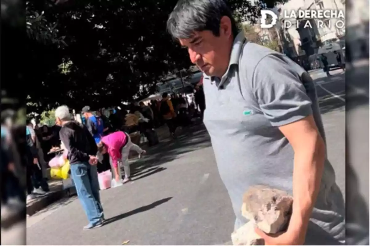 Un hombre con una camiseta gris sostiene un par de piedras en una calle concurrida, mientras otras personas están ocupadas en el fondo.