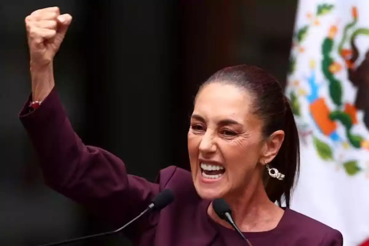 Una persona con expresión apasionada levanta el puño mientras habla frente a un micrófono con una bandera de México de fondo.