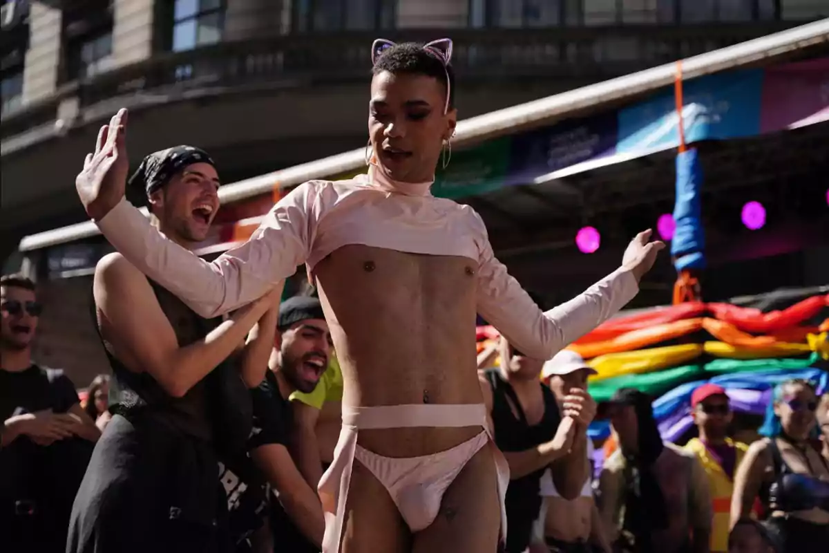 Persona con orejas de gato y atuendo rosa bailando en un evento al aire libre con una bandera arcoíris de fondo.