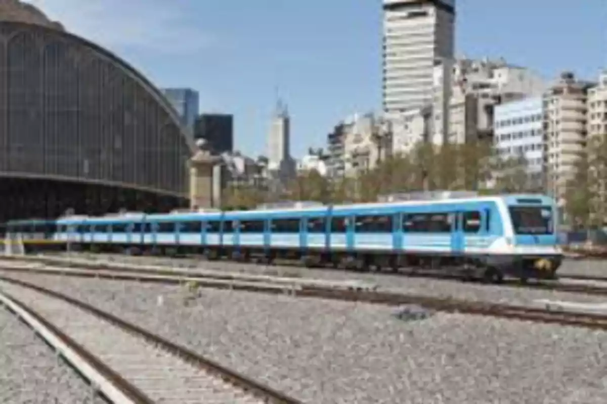 Un tren azul y blanco en una estación con edificios de fondo en un día soleado.