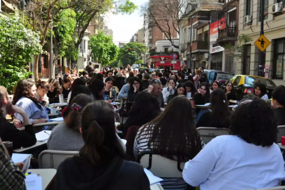 Un grupo numeroso de personas sentadas en escritorios al aire libre en una calle, rodeados de edificios y árboles.