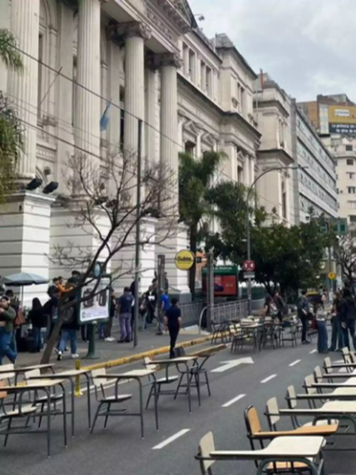 Personas reunidas frente a un edificio con columnas clásicas y escritorios en la calle.