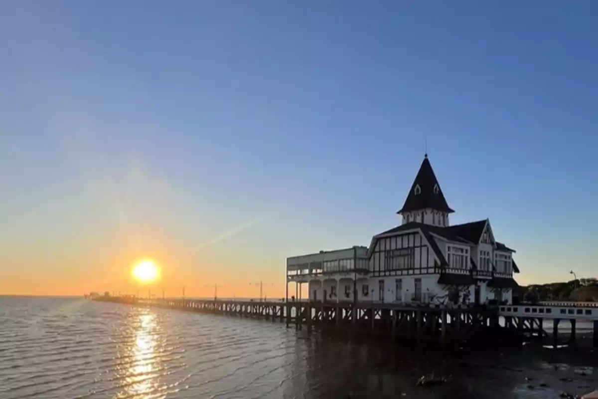 Atardecer sobre un muelle con un edificio de estilo clásico y el sol reflejándose en el agua.