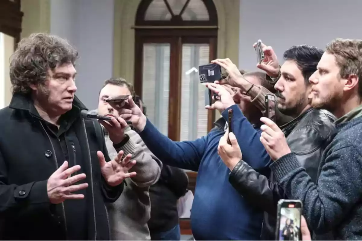 Un hombre hablando mientras varias personas lo graban con sus teléfonos en una sala con ventanas grandes.