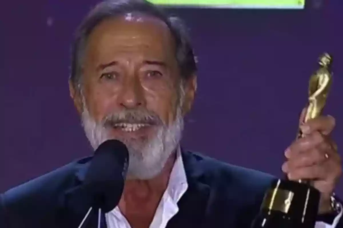 Un hombre con barba y cabello canoso sostiene un trofeo dorado mientras habla frente a un micrófono.