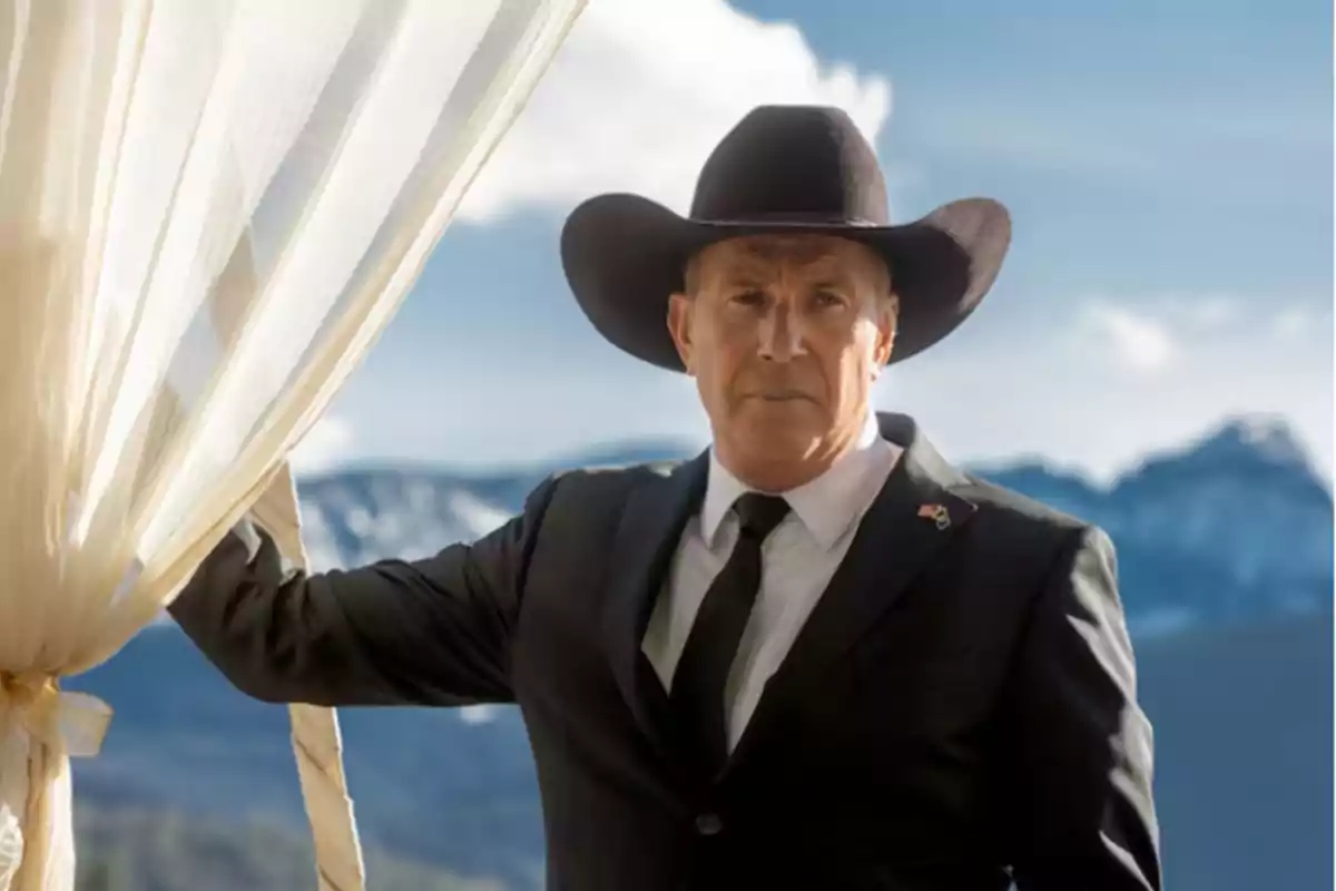 Hombre con sombrero de vaquero y traje oscuro, de pie junto a una cortina blanca con montañas nevadas al fondo.