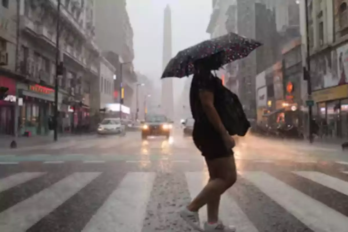 Persona cruzando una calle bajo la lluvia con un paraguas en una ciudad con edificios y tráfico.