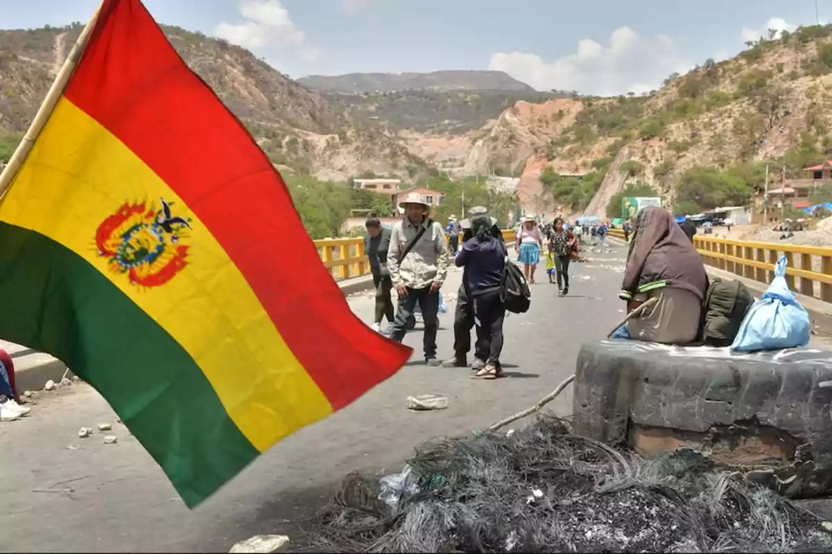 Una bandera de Bolivia ondea en un puente mientras personas caminan y se sientan cerca de un neumático en un entorno montañoso.