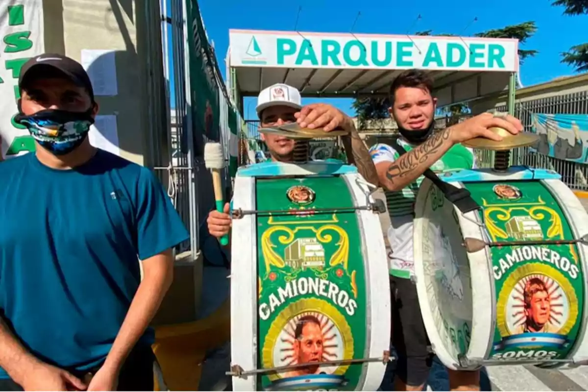 Tres personas posan con tambores decorados con el logo de "Camioneros" frente a un cartel que dice "Parque Ader".