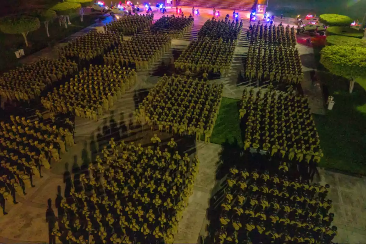 Una formación nocturna de soldados en un patio iluminado con luces de vehículos al fondo.