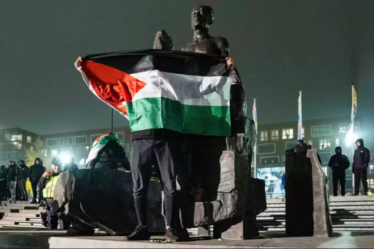 Una persona sostiene una bandera palestina frente a una estatua durante la noche, rodeada de otras personas y luces de edificios al fondo.