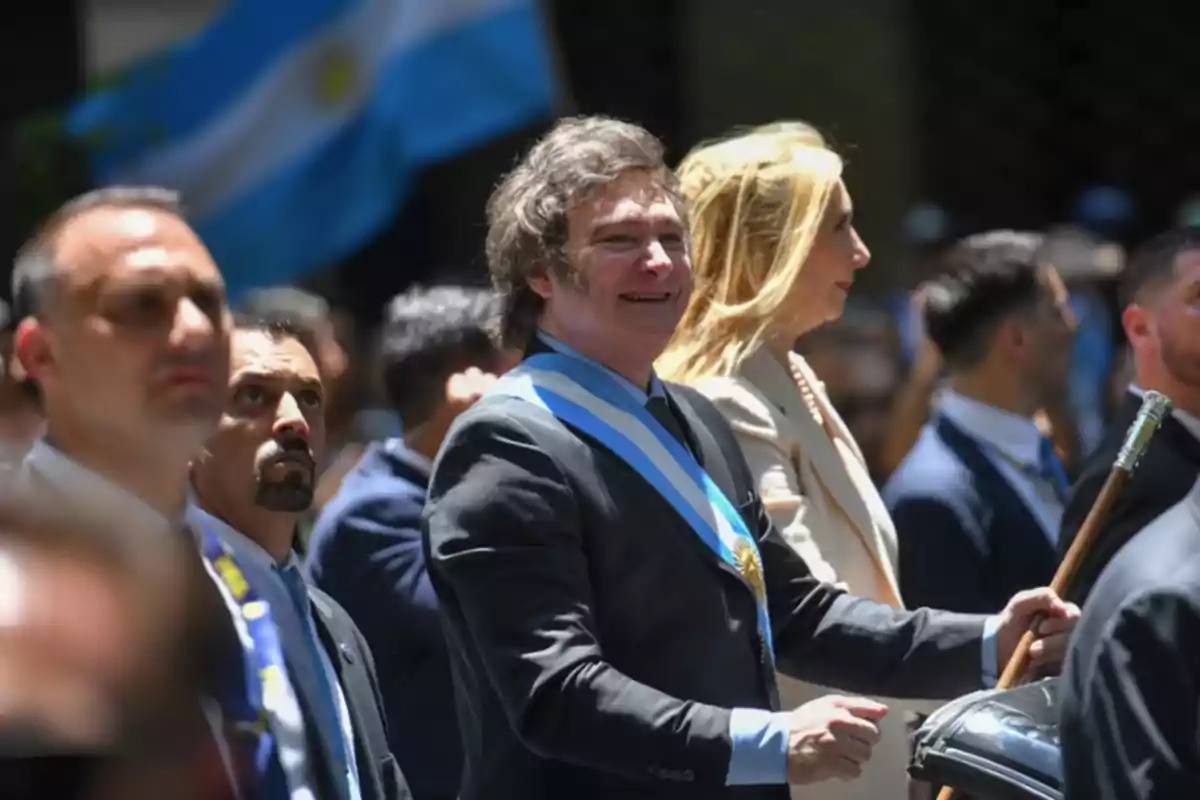 Un grupo de personas vestidas formalmente camina al aire libre con una bandera argentina de fondo.