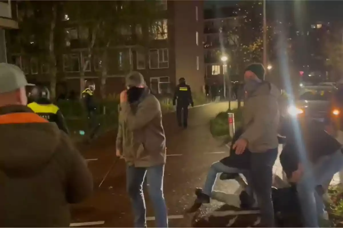 Un grupo de personas en una calle por la noche con presencia policial al fondo.