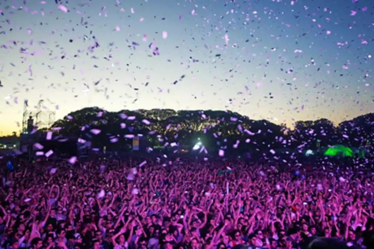 Una multitud disfrutando de un concierto al aire libre con confeti volando en el aire al atardecer.