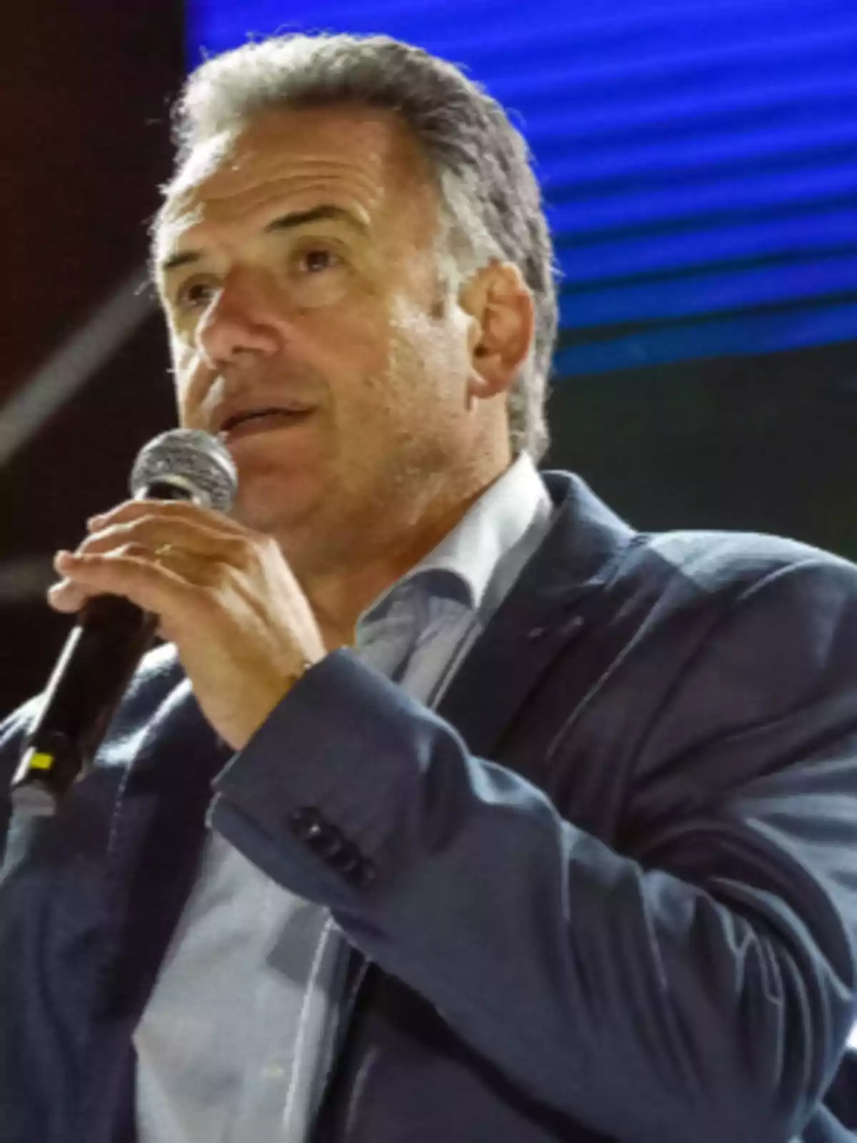 Hombre de cabello canoso hablando por micrófono en un evento con fondo azul.