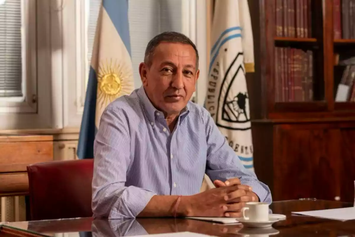 Hombre sentado en una oficina con banderas y estanterías de libros al fondo.